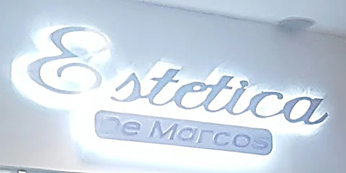 El letrero del local: “Estética” en letra cursiva cromada sobre una placa que dice “De Marcos”, con luz azul detrás.